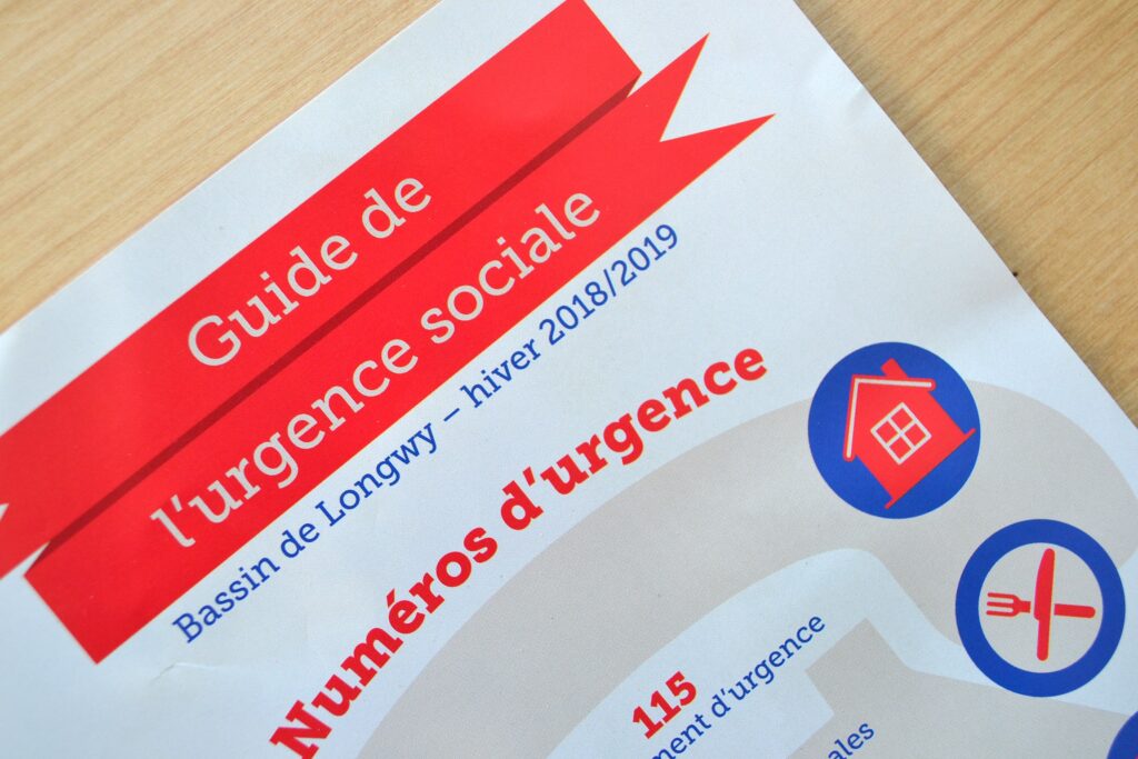 couverture du Guide de l'urgence sociale : titre dans une bannière rouge en haut au centre de la page. Sur un fond beige se détache un téléphone beige un peu plus foncé. Par dessus on lit les numéros d'ugence, accompagnés d'un pictogramme qui les représente.