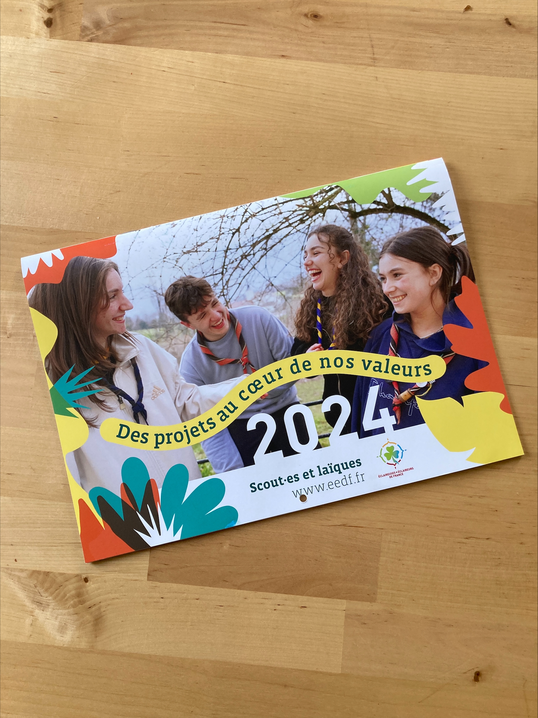 Couverture du calendrier : sur la photo en pleine page, trois filles et un garçons rigolent. Il est écrit : "Des projets au cœur de nos valeurs" et l'année, 2024. sur les bords de la page, il y a des fleurs. 